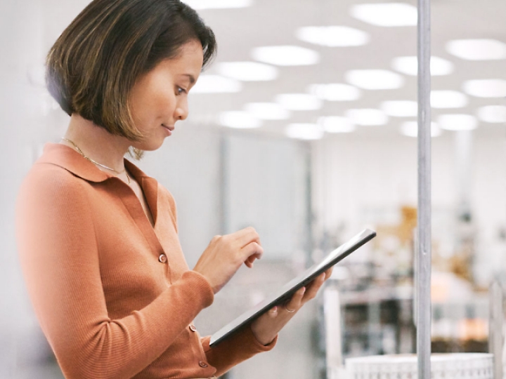 Une femme en haut orange utilise une tablette tout en se tenant dans une pièce bien éclairée, éventuellement un bureau ou un labo.