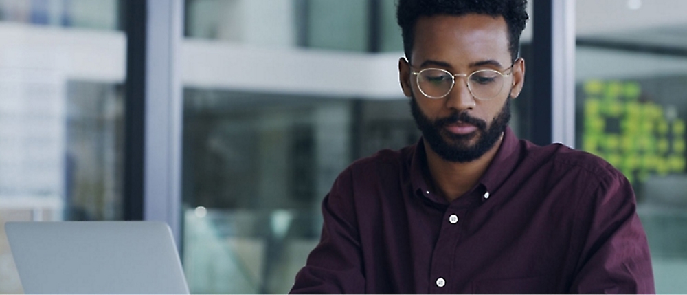 A person with glasses and a beard is sitting at a desk, focused on their laptop. A glass window is in the background.