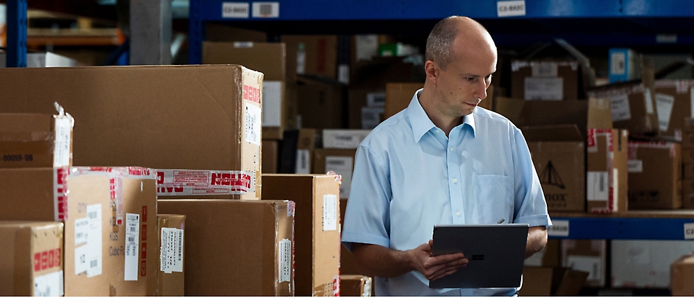 A man in a light blue shirt stands in a warehouse filled with cardboard boxes, holding a tablet.