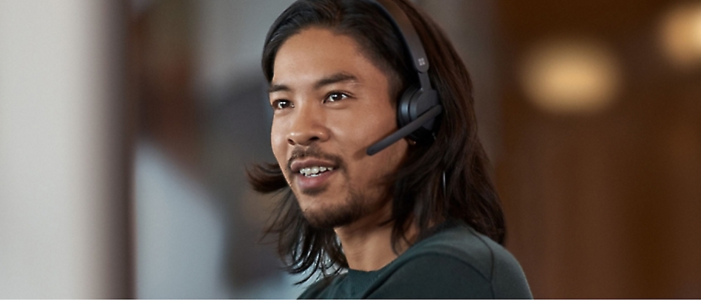 A person with long hair wearing a headset and speaking, with a blurred background.