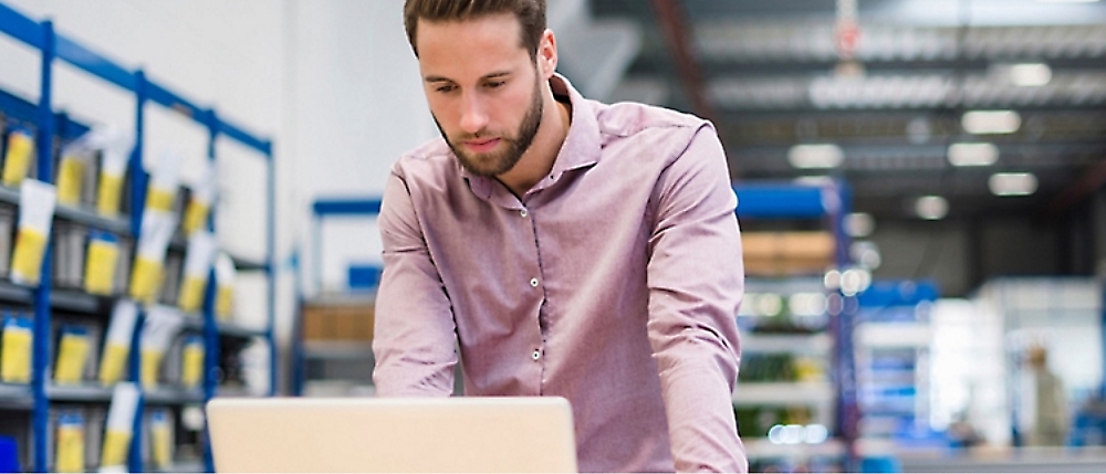 A man in a purple shirt works on a laptop in a warehouse with shelves lined with boxes.