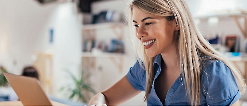 A woman with long blonde hair smiles while working on a laptop in a bright room. 