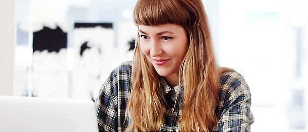 A person with long hair and bangs, wearing a plaid shirt, is looking at a laptop screen and smiling.