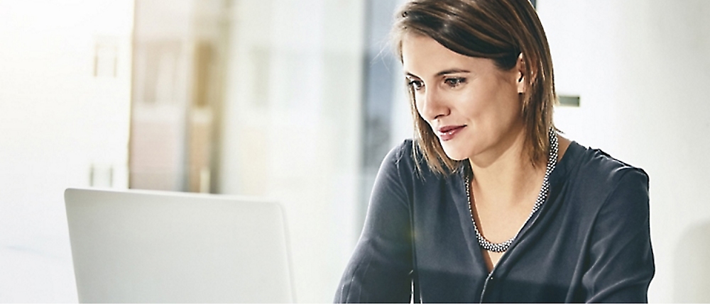 A woman with shoulder-length hair and wearing a dark blouse is sitting at a desk, looking at a laptop screen.