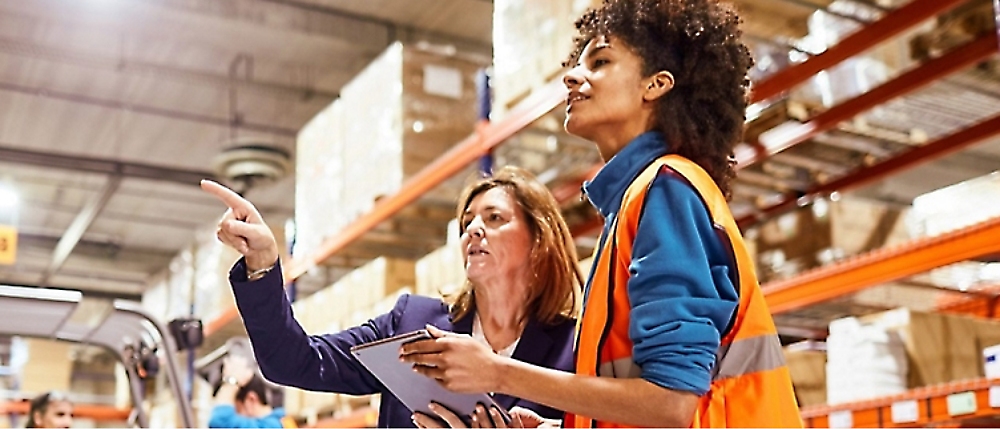 Two women in a warehouse, one in a business suit holding a tablet