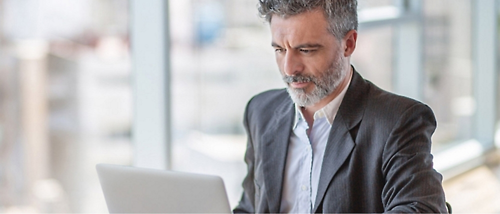 A man with grey hair and a beard works on a laptop computer in a bright, modern office setting.