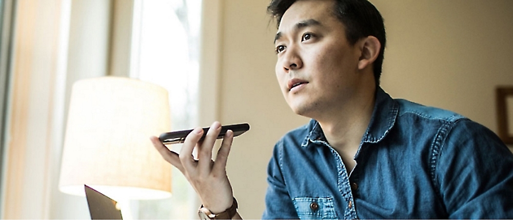 A man in a denim shirt holds a smartphone to his mouth, appearing to speak or record a message. 