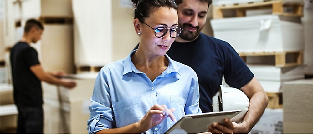 A woman in glasses uses a tablet while a man holding a helmet stands behind her in a warehouse. 