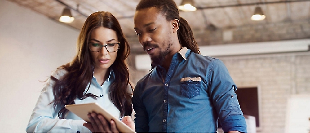 Two people in casual office attire collaborate while looking at a tablet. 