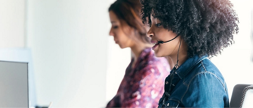 Two women sit in an office working with headsets on, focusing on their computer screens. 