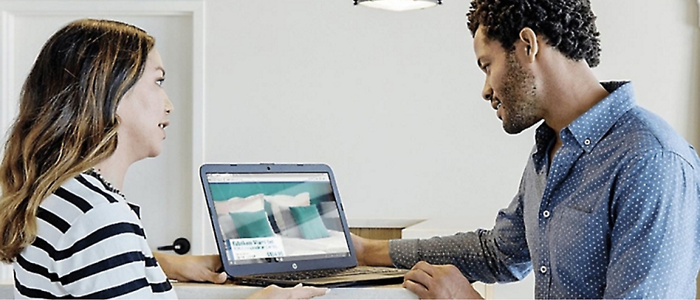 Two people are looking at a laptop screen displaying an image of a bed with pillows in a room setting.