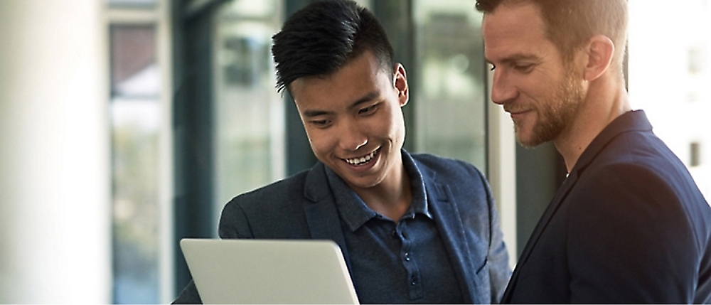 Two men in business attire look at a laptop screen in an office setting, one smiling and the other observing