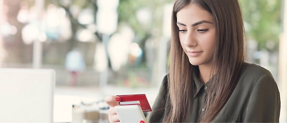 A woman is sitting indoors, holding a smartphone in one hand and a credit card in the other