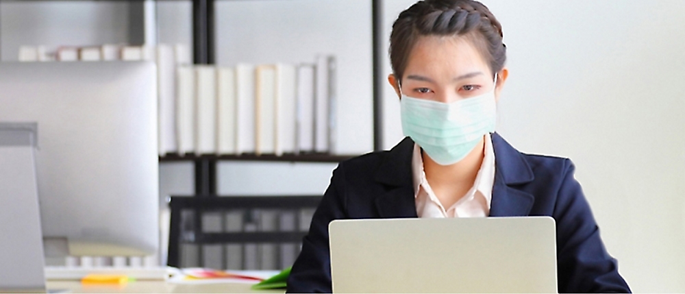 A person wearing a face mask works on a laptop in an office setting with shelves and files in the background.