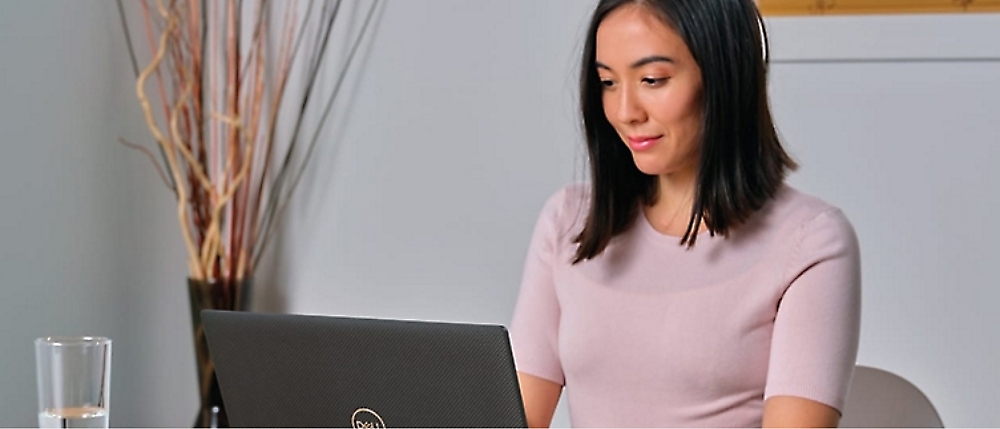 A woman in a light pink top is working on a Dell laptop at a desk with a glass of water.