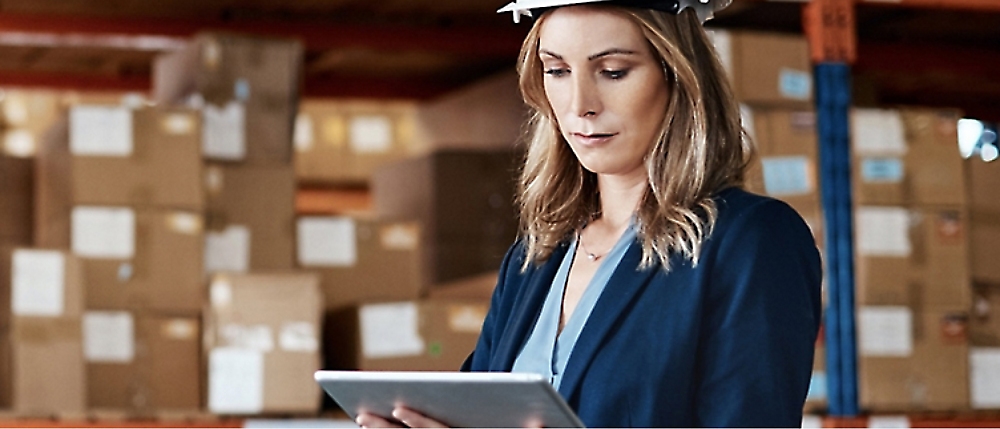 A person wearing a hard hat and blazer is using a tablet in a warehouse filled with stacked boxes.