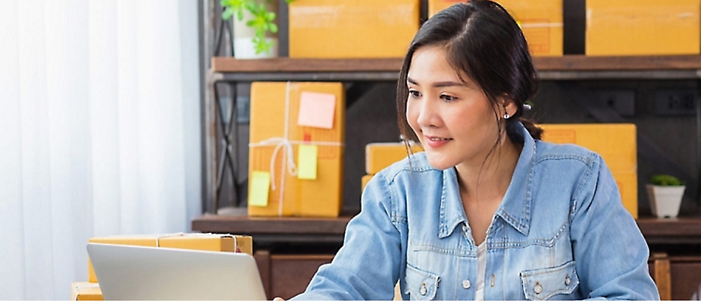 A woman in a denim shirt uses a laptop at a desk cluttered with cardboard boxes.