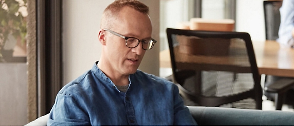 Man with glasses and short hair wearing a denim shirt, sitting on a couch and looking down. 