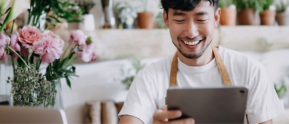 A person wearing an apron smiles while looking at a tablet. They are seated in a room filled with plants and flowers.