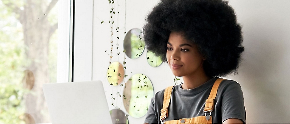 A person with a large afro hairstyle, wearing a grey shirt and yellow overalls, looks at a laptop 