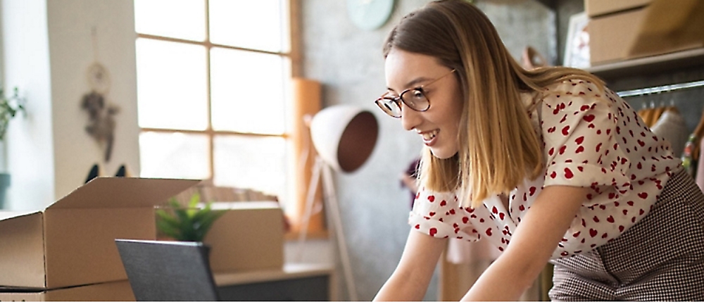 A woman wearing glasses and a white shirt with red hearts works at a laptop in a room with cardboard boxes and shelves.