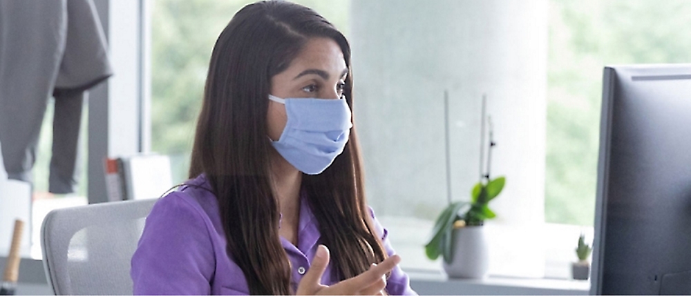 A woman wearing a face mask sits at a desk, gesturing with her hands while speaking. 