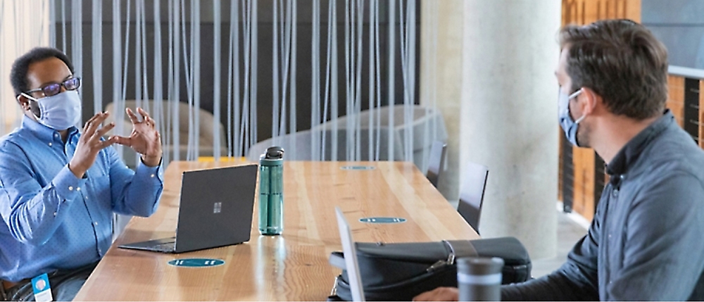 Two individuals, both wearing masks, engage in a discussion across a conference table with laptops 