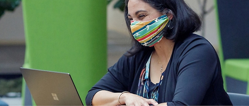 A woman wearing a colorful face mask works on a laptop.