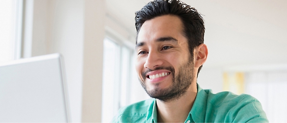 A person with dark hair and a beard, wearing a green shirt, is smiling while looking at a computer screen.