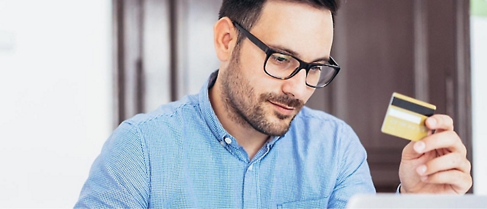 A man wearing glasses and a blue shirt holds a credit card while looking at a laptop.