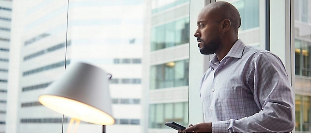 A man in a checkered shirt stands by a window in an office building, looking outside while holding a phone.