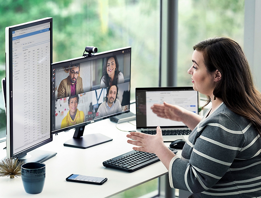 A woman sits at a desk in front of multiple computer screens, participating in a video conference call with four other people