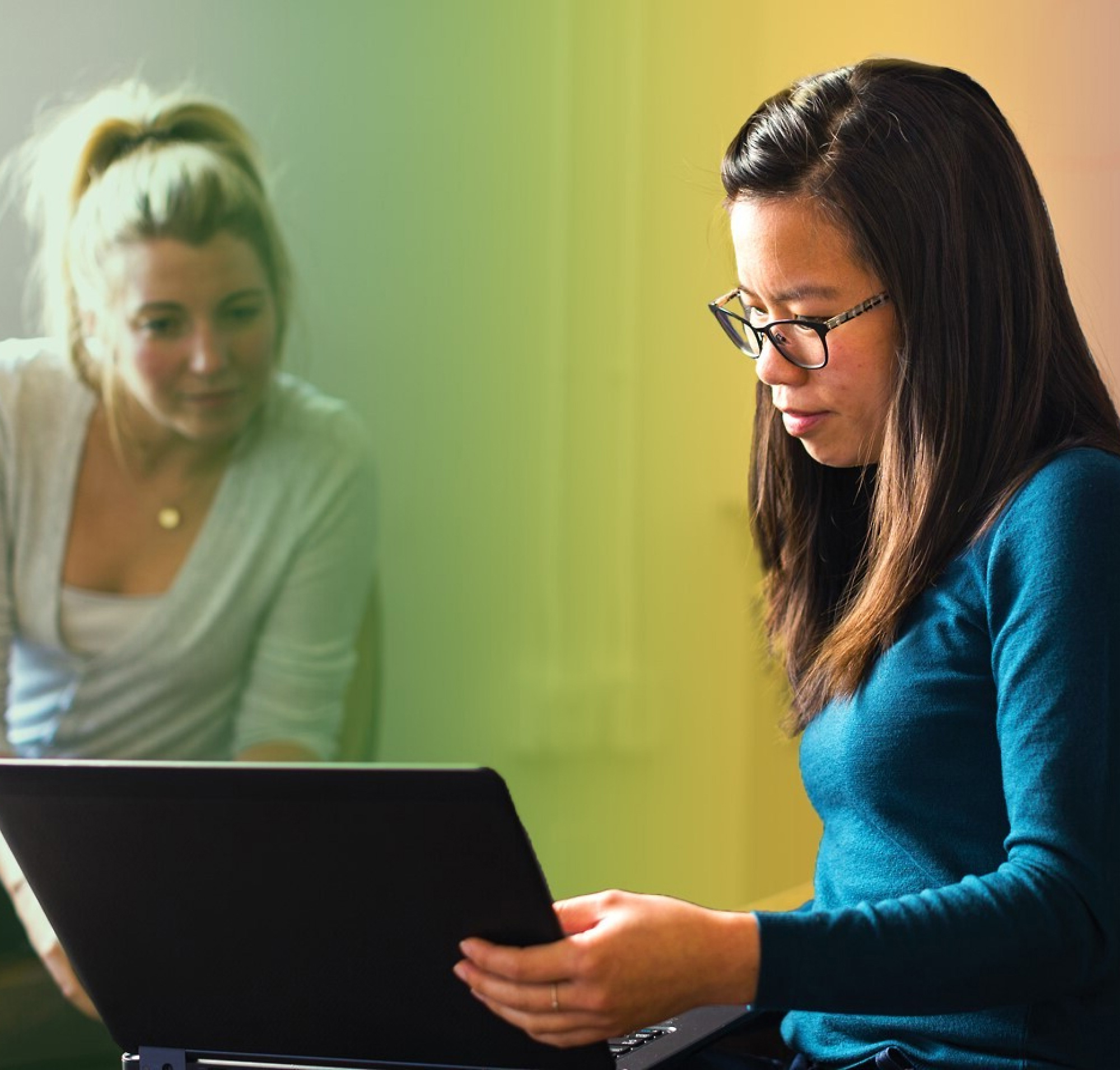 Twee vrouwen, de een met blond haar en de ander met een bril, werken samen op een laptop in een kleurrijk verlichte kamer.