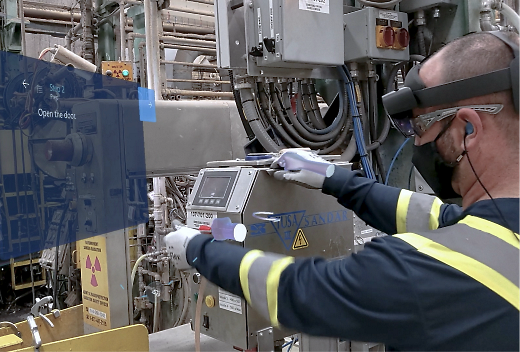 A worker in a safety vest and headset uses a control panel in an industrial setting with complex machinery