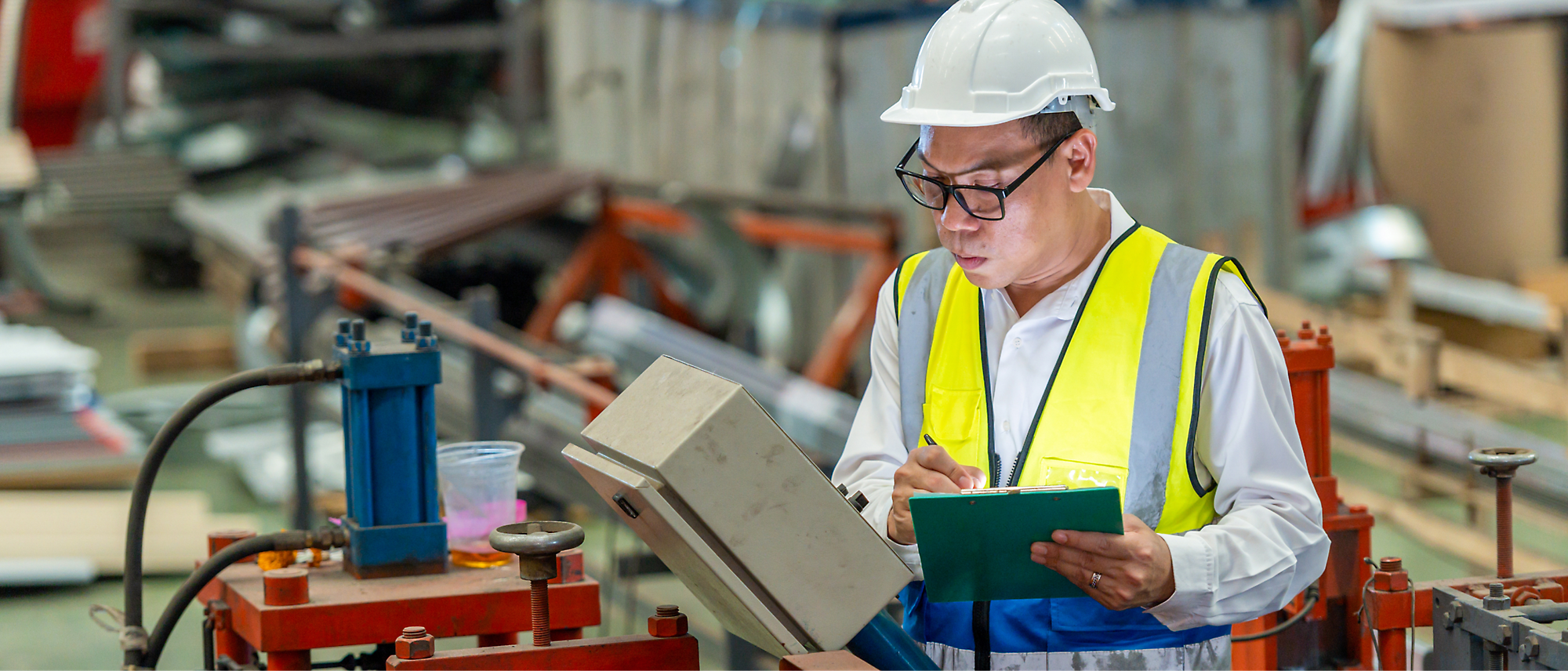 Seorang pria dengan topi pengaman dan rompi visibilitas tinggi menulis di clipboard saat memeriksa mesin dalam area industri.