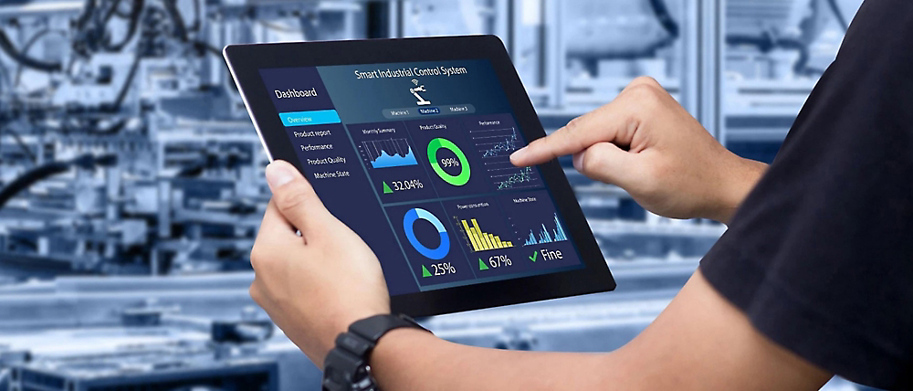 En person som bruker et digitalt nettbrett som viser ulike analyser foran en uskarp industriell bakgrunn.