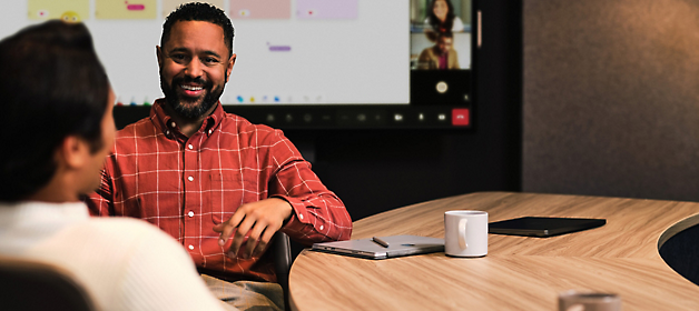 Two professionals discuss at a meeting table, with a large screen showing a video call in the background.