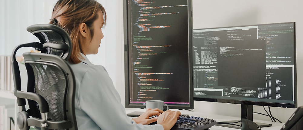 A person seated at a desk, typing on a keyboard while looking at code displayed on two large computer monitors.