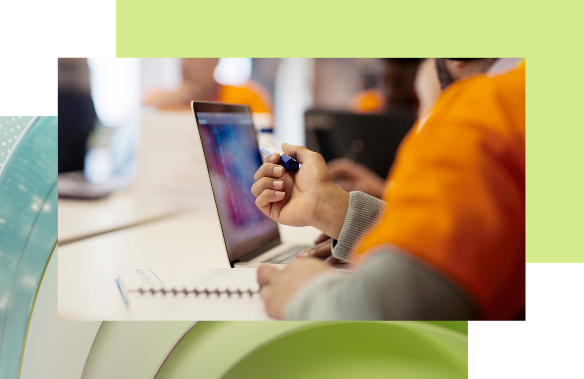 A person in an orange jacket using a stylus on a laptop screen at a busy workspace.