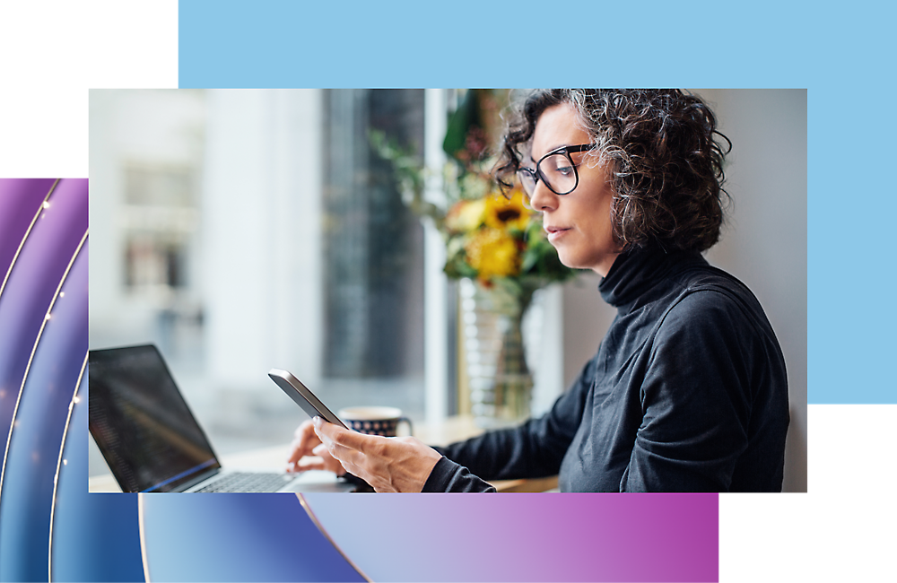 A woman with curly hair and glasses using a smartphone and laptop at a modern workspace.