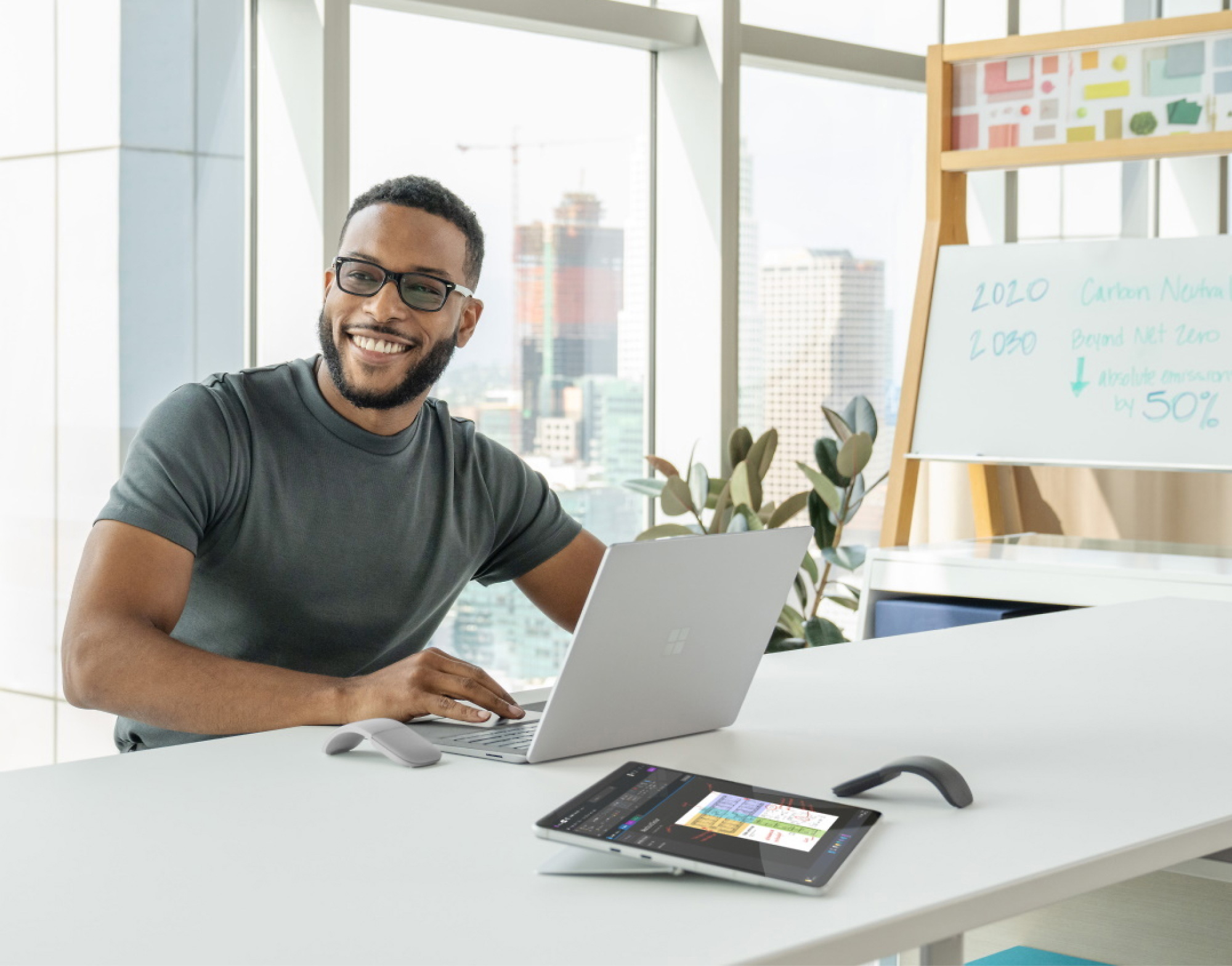 Homme souriant portant des lunettes et utilisant un ordinateur portable à un bureau moderne où se trouvent une tablette, un téléphone et un tableau de présentation