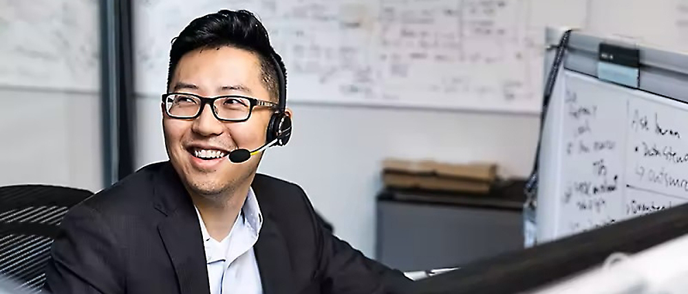 Een lachende man met een headset en bril kijkt naar een computerscherm in een kantoor met een whiteboard op de achtergrond.