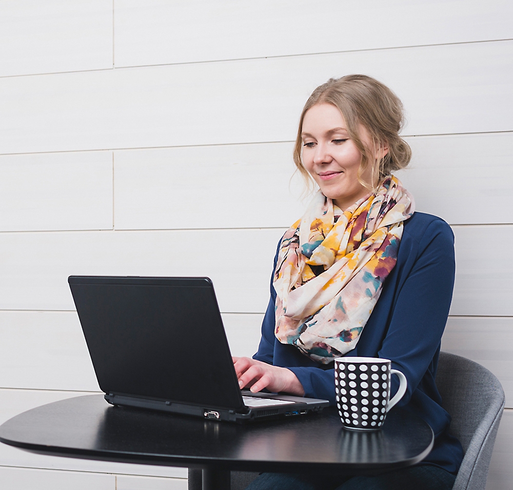 Uma mulher de suéter azul e cachecol colorido sorrindo enquanto usa um laptop em uma mesa com uma caneca de café ao lado.