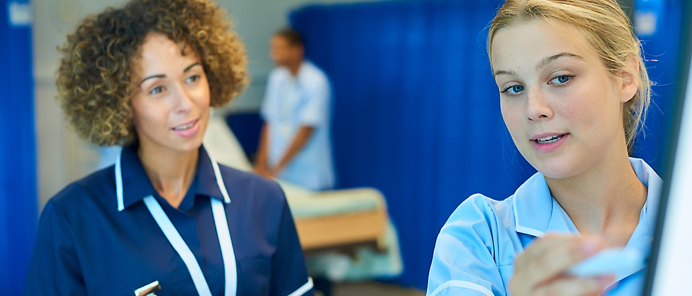 Two female healthcare professionals in a hospital, one pointing and explaining something, with another staff member 