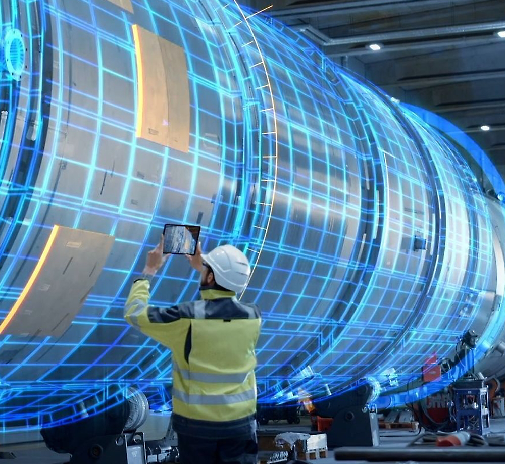 A person wearing a hard hat and high-visibility jacket stands in front of a large cylindrical structure, holding a tablet. 