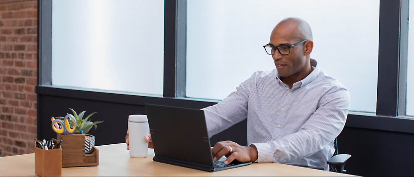 A person wearing glasses and a light-colored shirt sits at a desk, typing on a laptop 