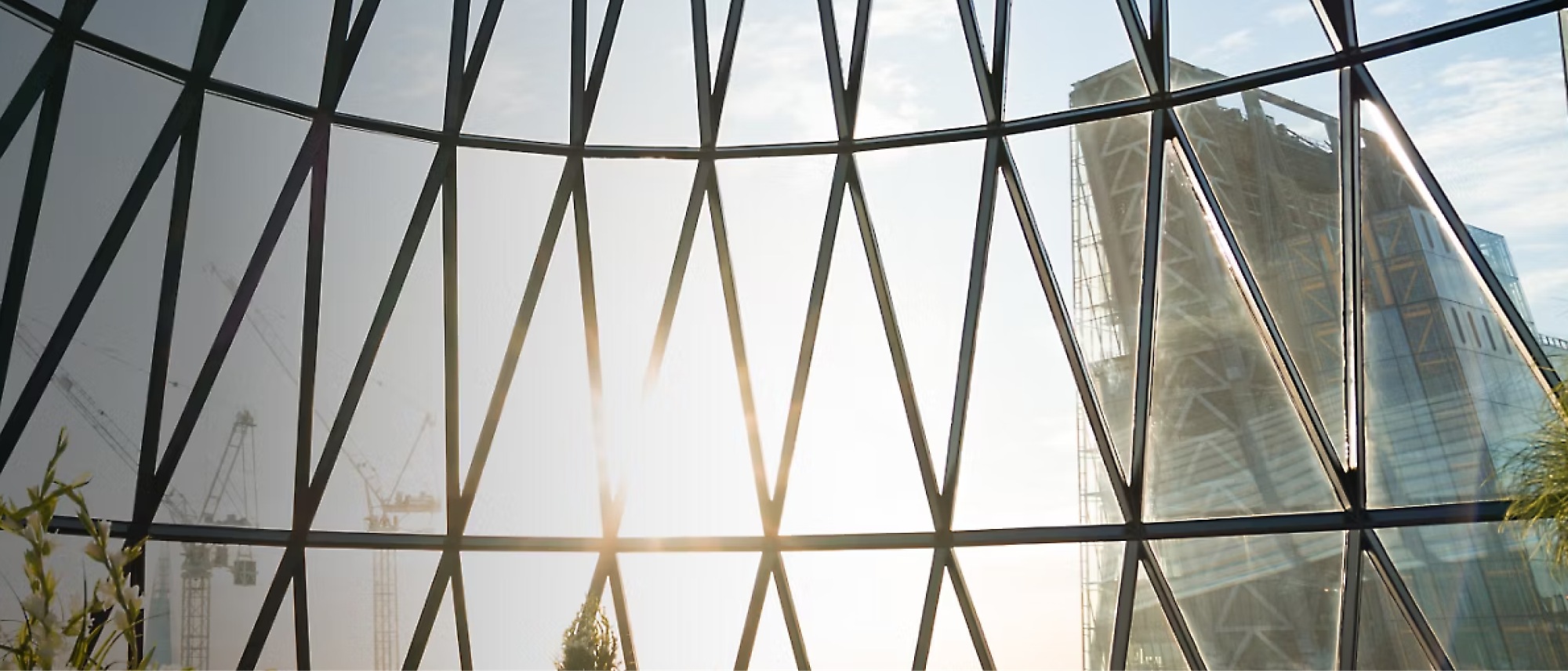 View through a grid-patterned window showcasing a modern glass building and several construction cranes with sunlight shining through.