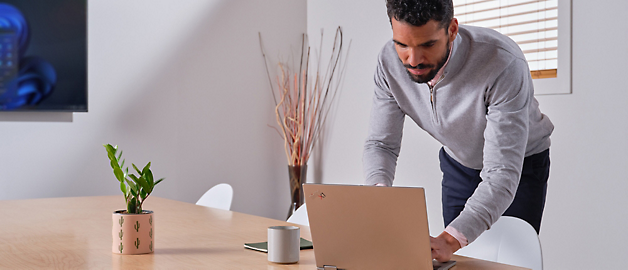 Mand i grå sweater står og læner sig ind over en bærbar computer på et træbord i et lyst kontor.