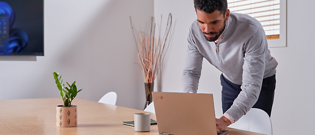 Man in gray sweater standing and leaning over a laptop on a wooden desk in a bright office.