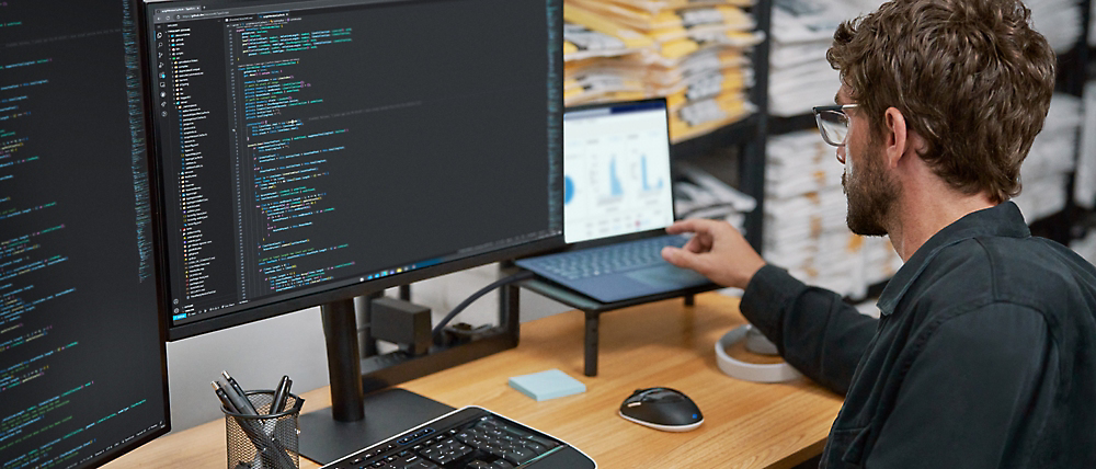 A man analyzing data on a computer with dual monitors displaying code and graphs in an office environment.